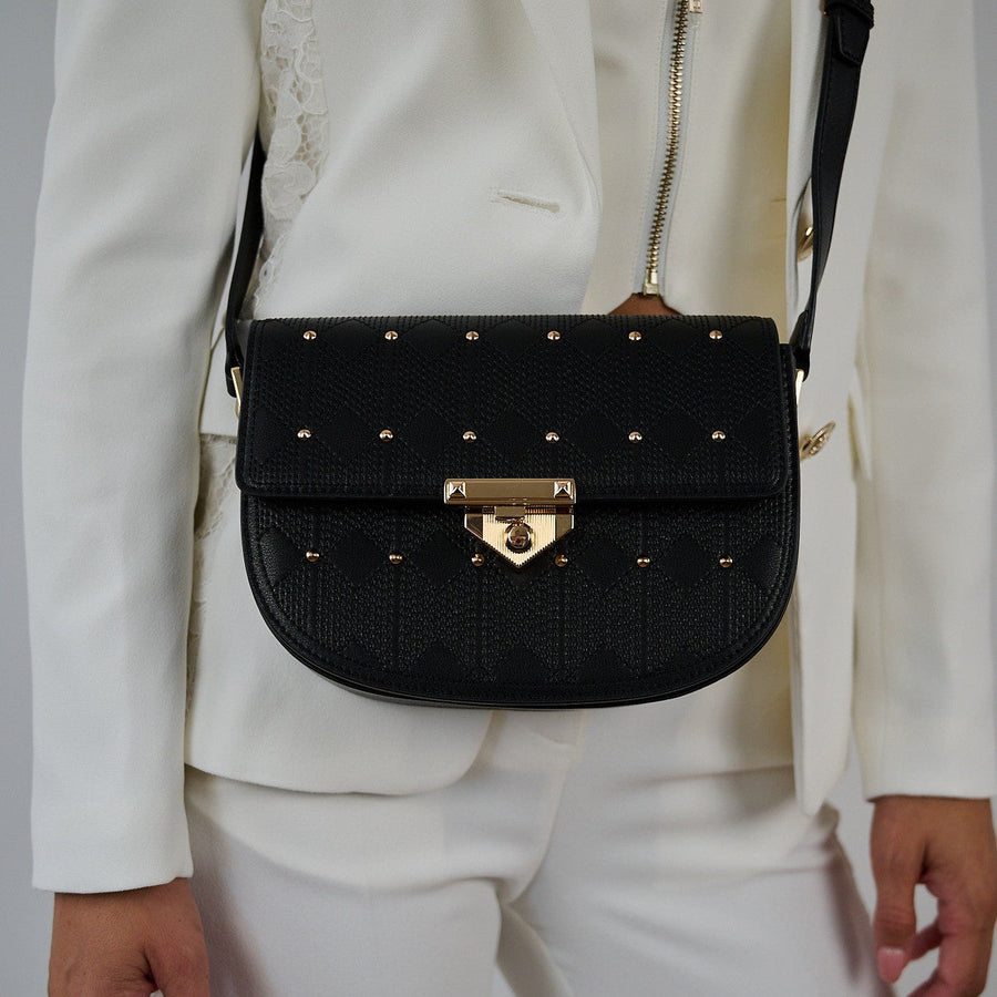 Naelie crossbody bag in black quilted Italian leather with gold studs, worn over a white suit